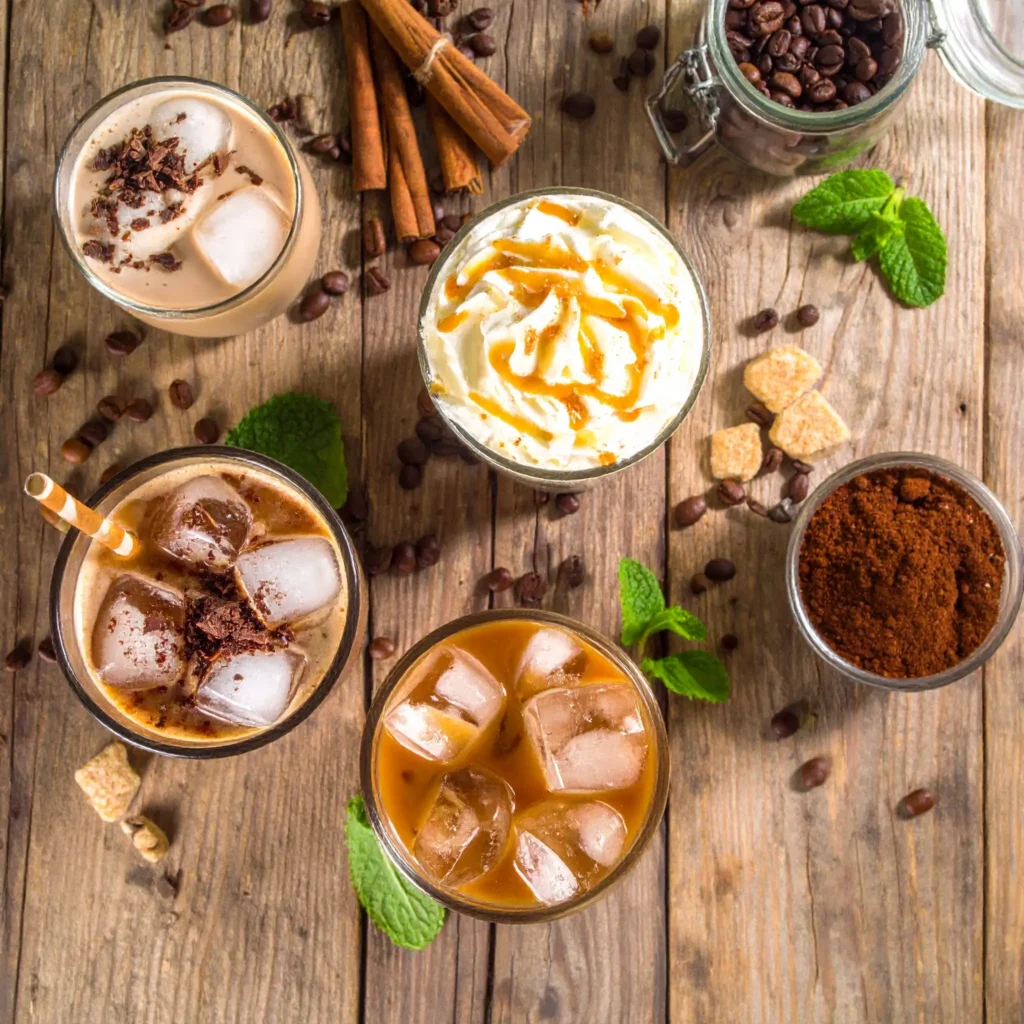 Flat lay of three iced coffee copycat drinks in clear glasses with whipped cream, caramel drizzle, and chocolate shavings, surrounded by coffee beans, mocha powder, cinnamon sticks, and mint on wooden table.