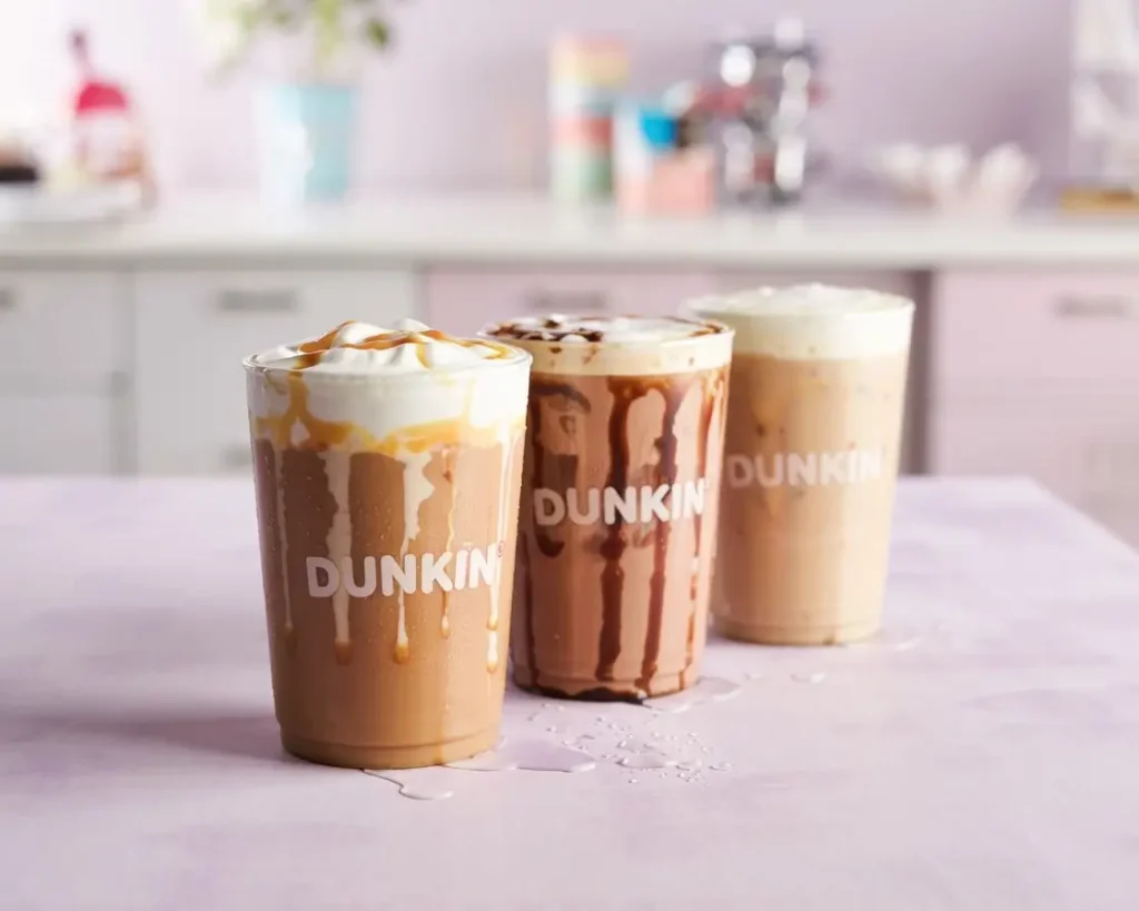 Three Dunkin iced coffee flavors in clear cups: caramel, mocha, and French vanilla on a pastel background.