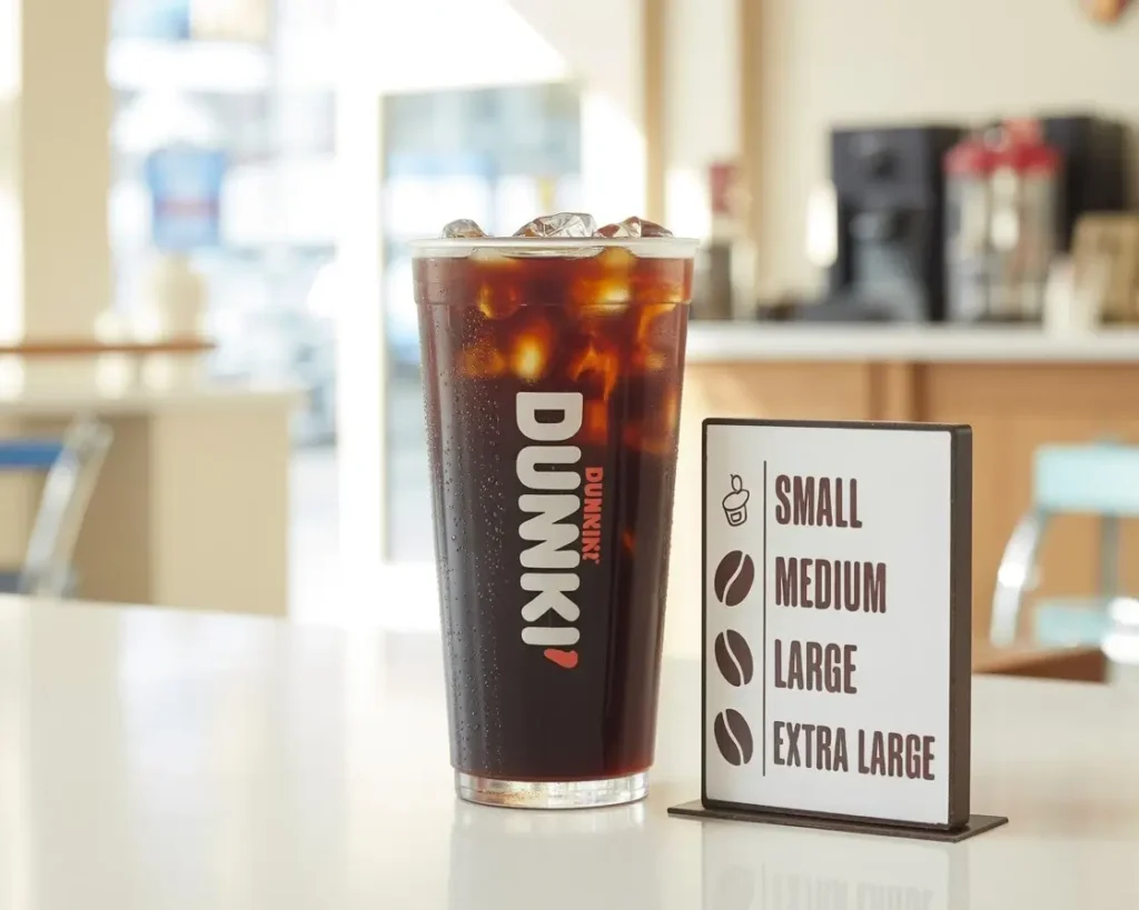 Dunkin’ iced coffee with ice beside a caffeine level chart showing different sizes on a café counter.