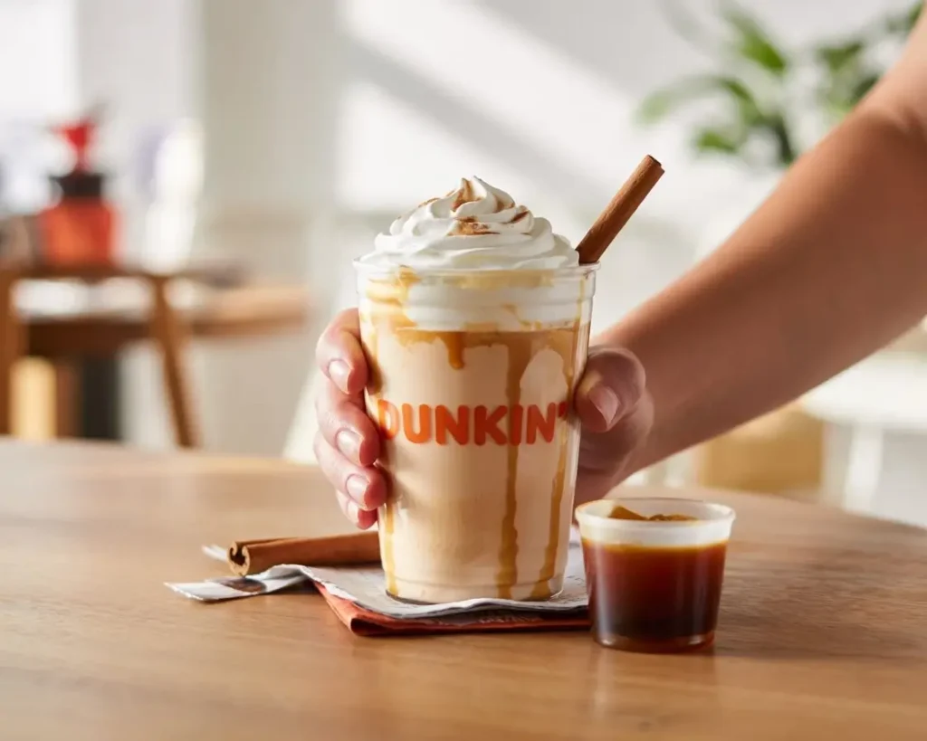 Hand holding Dunkin pumpkin spice latte with whipped cream and customization options on a wooden café table.