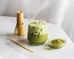 Iced matcha latte in a clear glass with ice, matcha powder, and a bamboo whisk on a marble surface.
